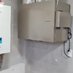 Zamontowany rekuperator Thessla Green z trzema srebrnymi, elastycznymi przewodami wentylacyjnymi w pomieszczeniu z szarymi płytkami, widoczny biały piec po lewej stronie.