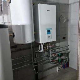 Biały zasobnik ciepłej wody użytkowej Galmet i wiszący dwufunkcyjny piec gazowy Panasonic z podłączeniami hydraulicznymi na tle szarej ściany z płytek ceramicznych.