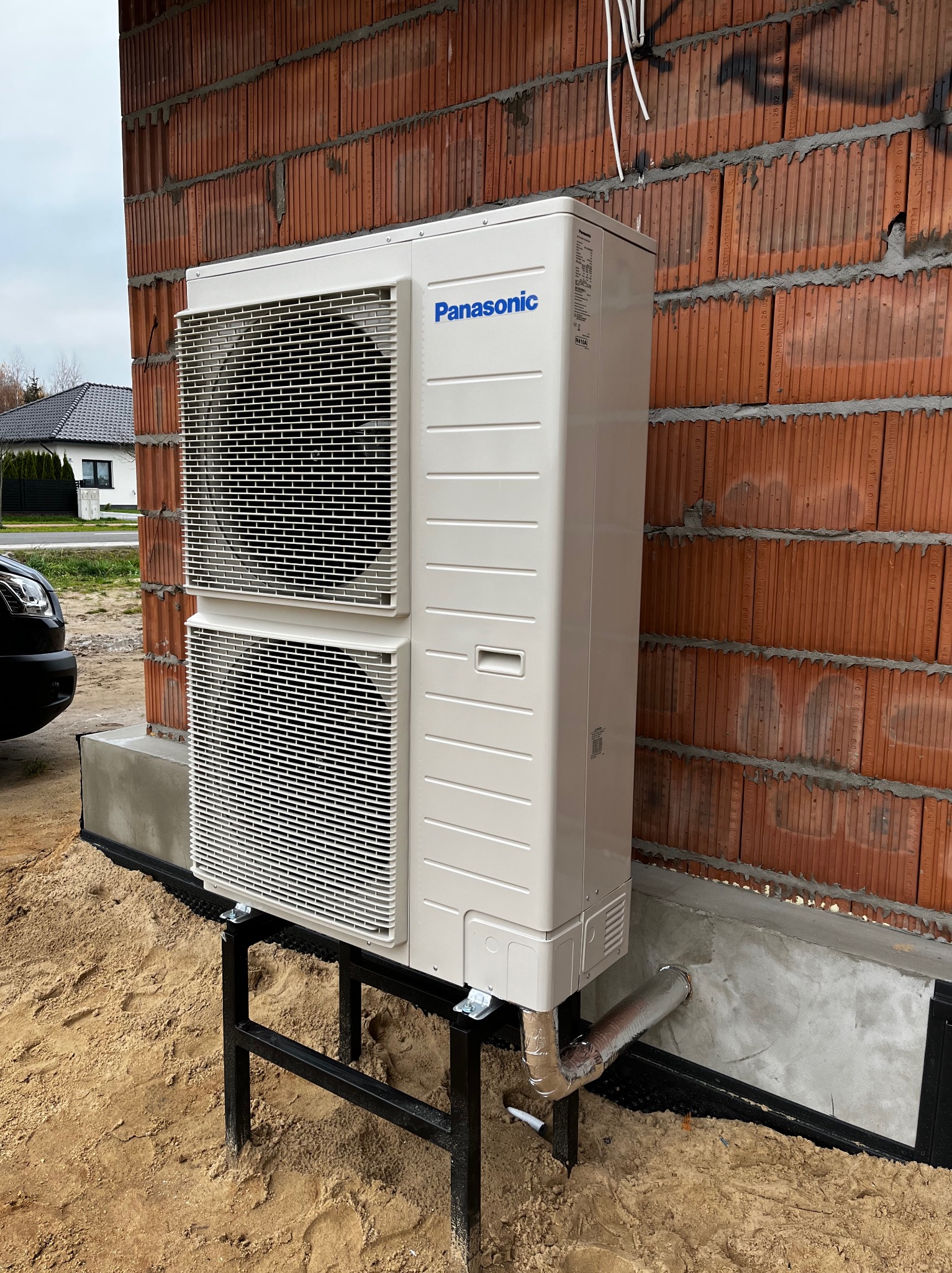 Zewnętrzna jednostka pompy ciepła Panasonic zamontowana na czarnej, stalowej konstrukcji przy ścianie budynku z czerwonej cegły kratówki, widoczny fragment instalacji odprowadzającej skropliny.
