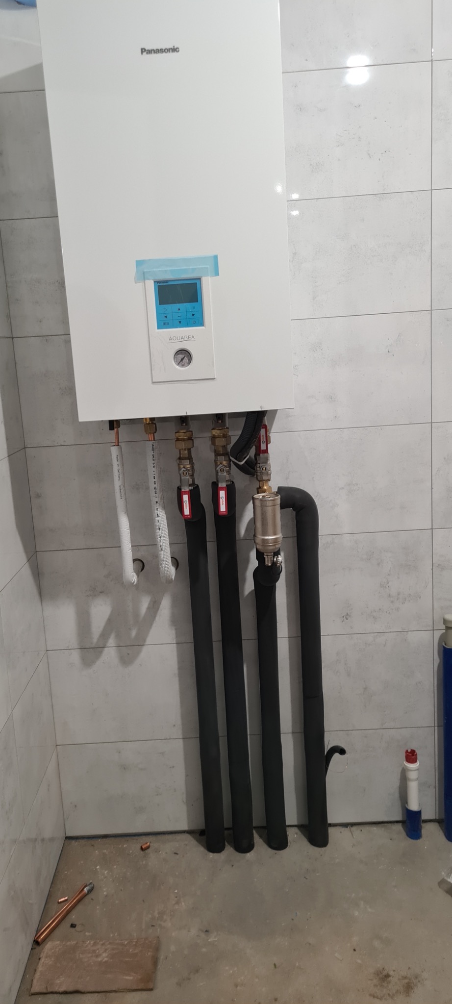Biała jednostka wewnętrzna Panasonic Aquarea z podłączonymi rurami grzewczymi w izolacji, zamontowana na ścianie z szarych płytek, widoczny filtr na jednej z rur.