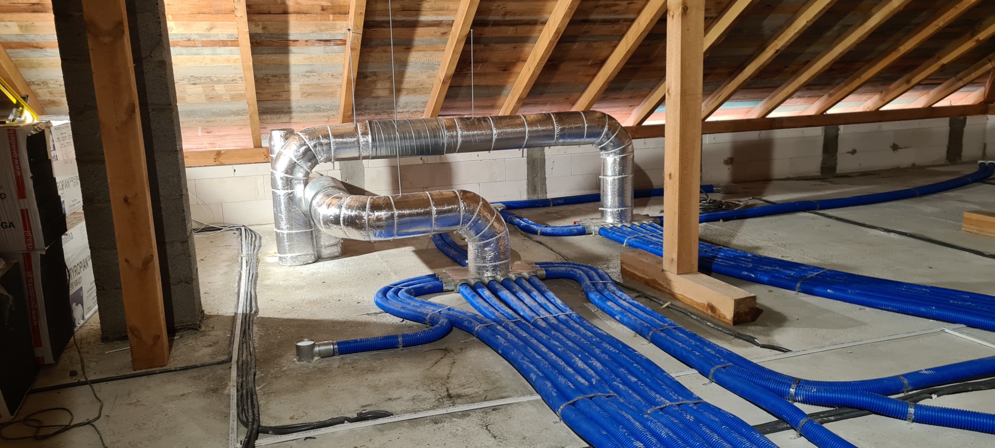 Instalacja przewodów wentylacyjnych z niebieskiego tworzywa sztucznego i metalowych rur w surowym wnętrzu poddasza z drewnianą konstrukcją dachu i ścianami z betonu komórkowego.