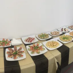 St&oacute;ł cateringowy z r&oacute;żnorodnymi przekąskami na białych talerzach, udekorowany czarno-beżowym obrusem w kratę i małymi etykietami informacyjnymi przy każdym talerzu, ustawiony pod białą ścianą.