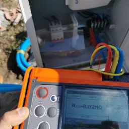 Pomiar rezystancji izolacji RISO 8,02MΩ na pomarańczowym mierniku elektrycznym, skrzynka z bezpiecznikami w tle, niebieskie rury instalacyjne obok.