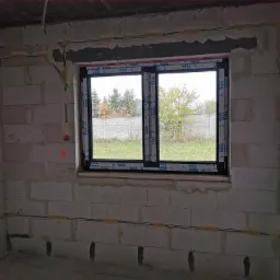 Surowy stan wnętrza z widocznymi przewodami elektrycznymi w ścianach z betonu komórkowego, okno z widokiem na ogród.