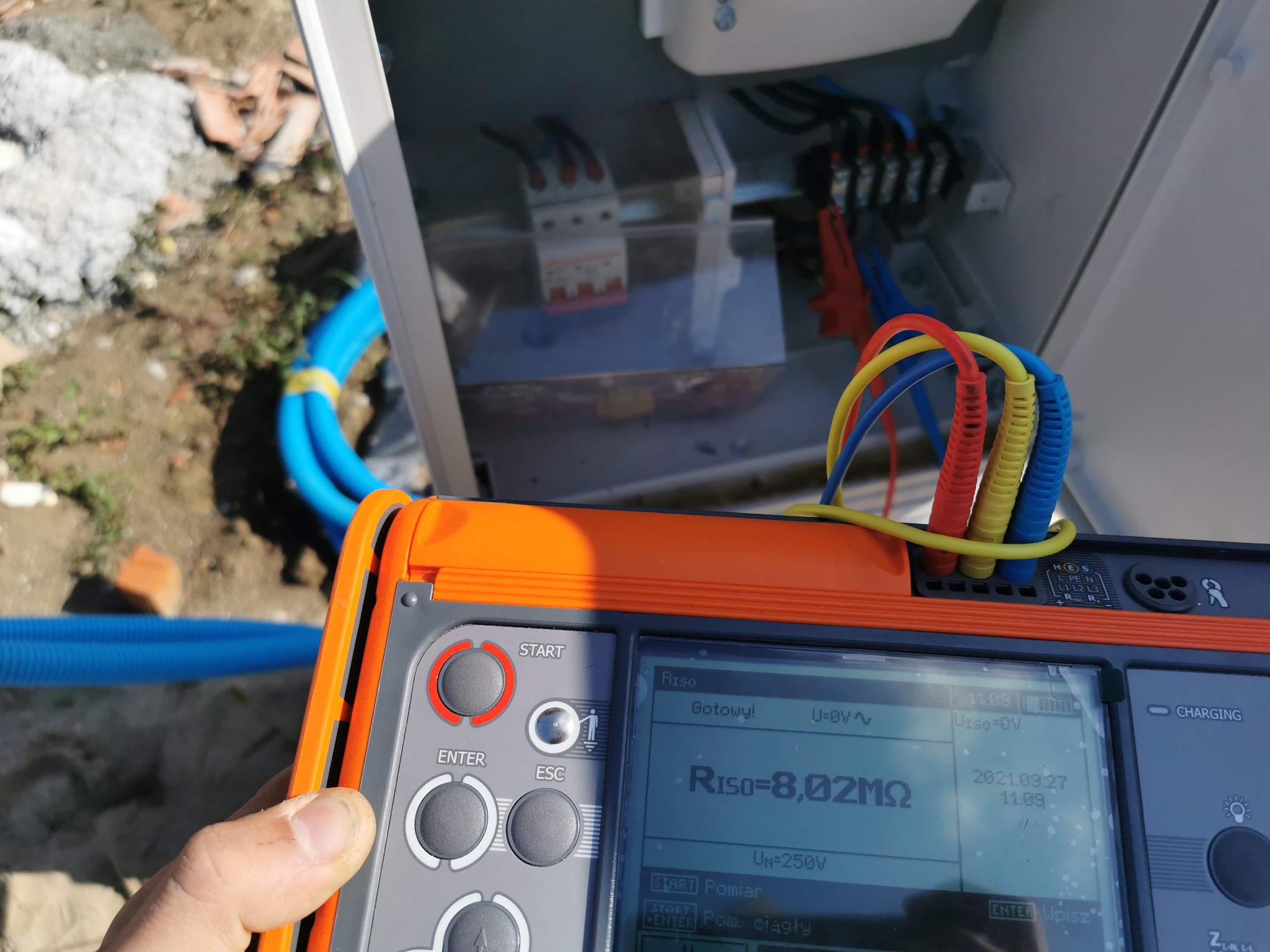 Pomiar rezystancji izolacji RISO 8,02MΩ na pomarańczowym mierniku elektrycznym, skrzynka z bezpiecznikami w tle, niebieskie rury instalacyjne obok.