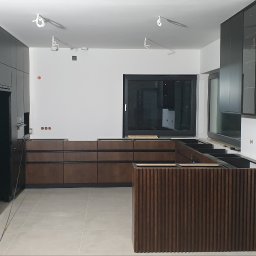 IMEB - Częściowo zmontowana kuchnia z czarnymi szafkami i drewnianymi frontami szuflad, widoczne przygotowane punkty elektryczne i oświetlenie sufitowe.