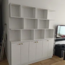 Biała, modułowa zabudowa meblowa z szafkami dolnymi i otwartymi półkami górnymi, z widoczną drabiną i fragmentem telewizora w tle.