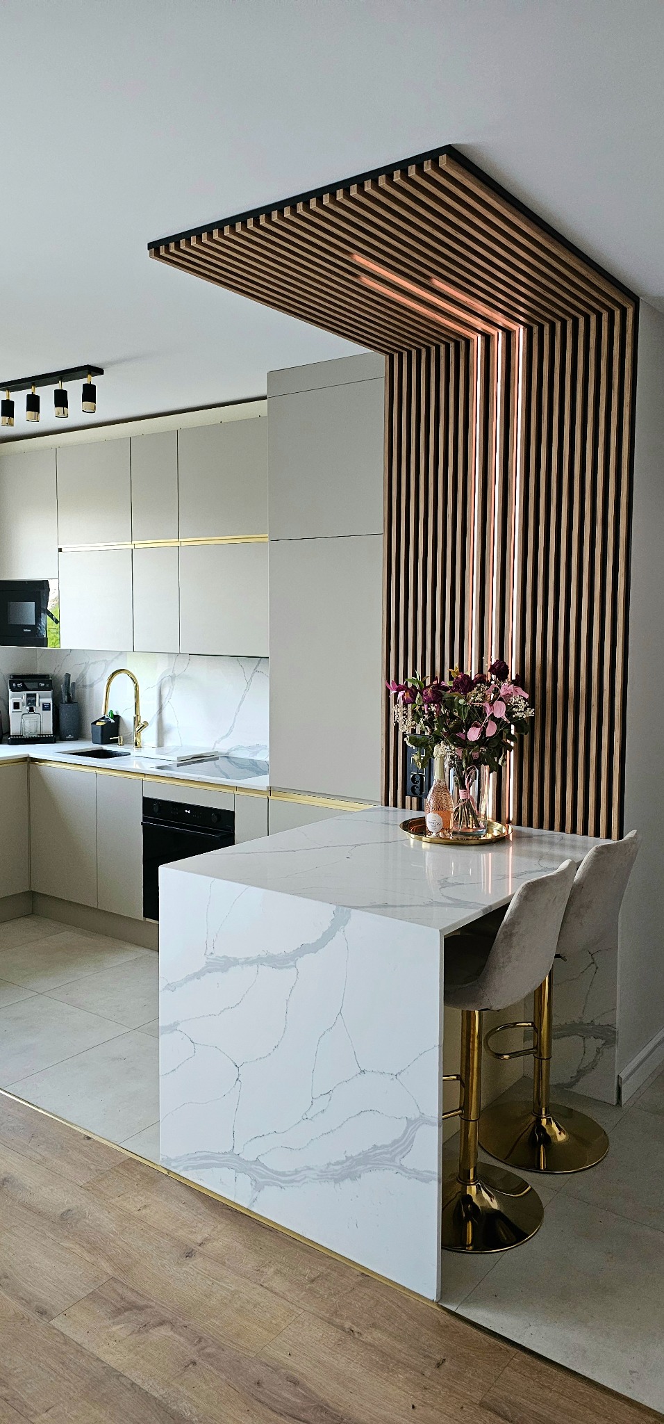Nowoczesna kuchnia z wyspą o marmurowym blacie, złotymi akcentami i drewnianą ścianką działową z podświetleniem LED.