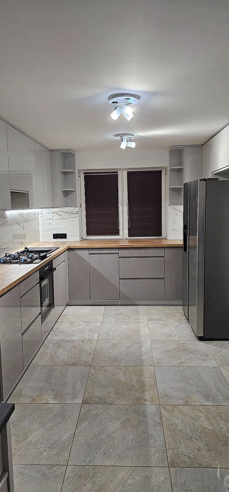 Nowoczesna kuchnia w odcieniach szarości z drewnianymi blatami, jasnymi płytkami imitującymi marmur na ścianie i szarą podłogą z płytek o wzorze kamienia.