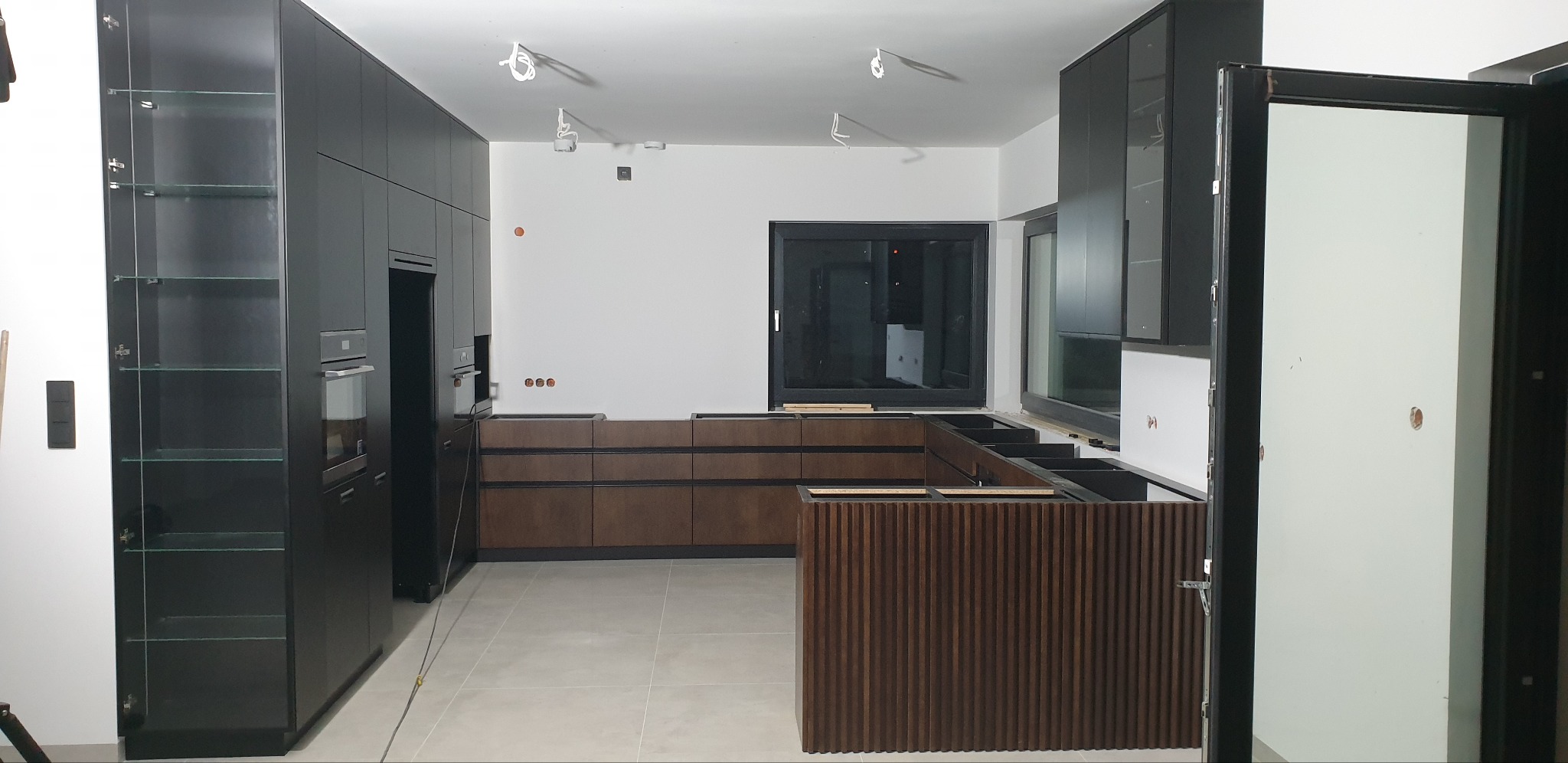 Częściowo zmontowana kuchnia z czarnymi szafkami i drewnianymi frontami szuflad, widoczne przygotowane punkty elektryczne i oświetlenie sufitowe.