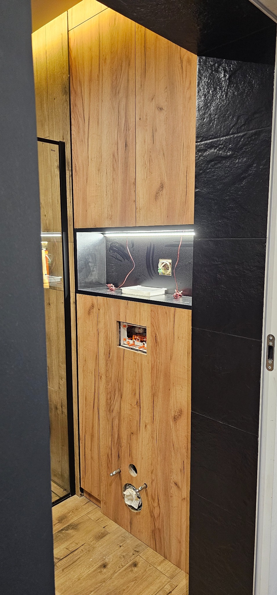 Montaż szafki z drewnopodobnymi frontami w łazience, widoczne podłączenia elektryczne i hydrauliczne w trakcie prac.