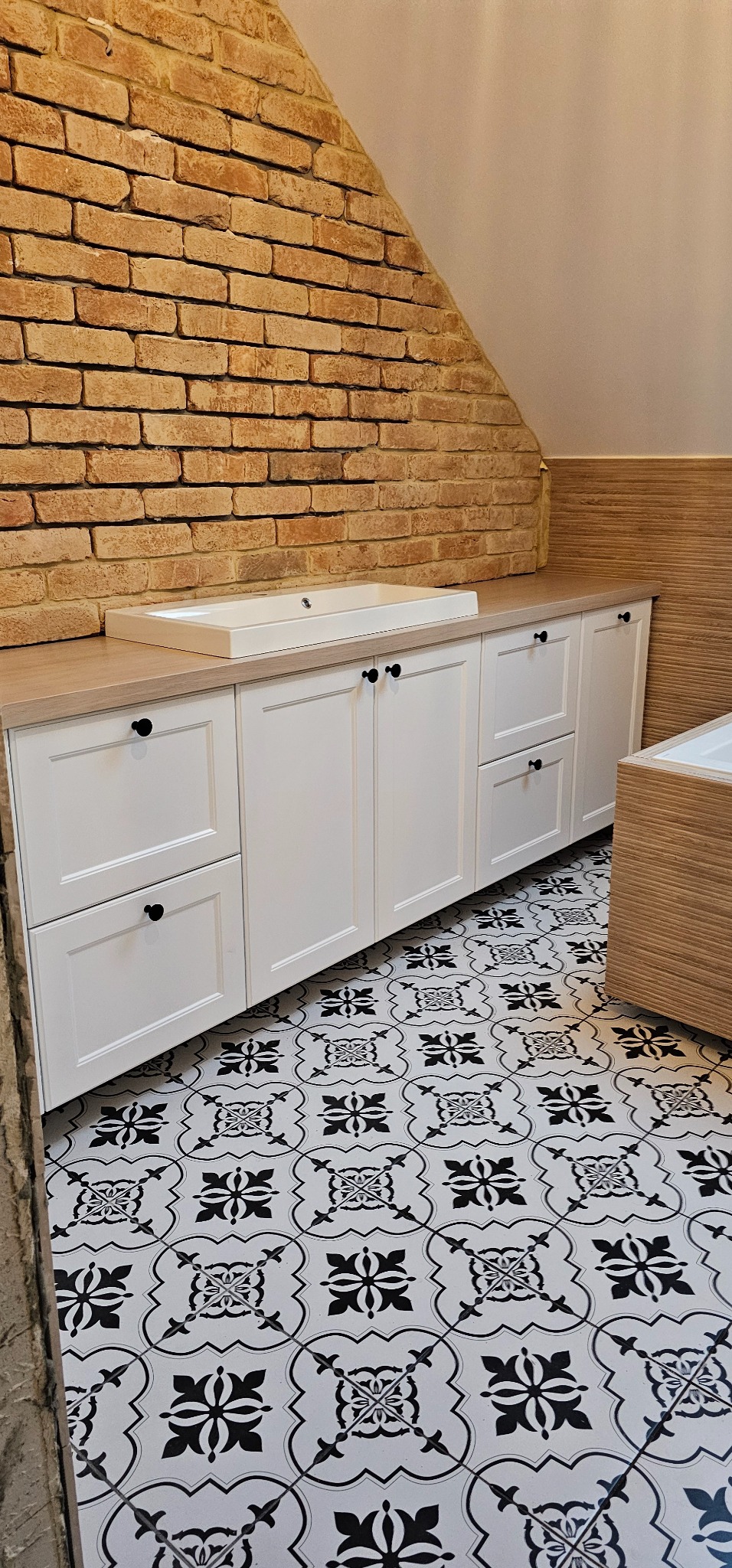 Białe szafki łazienkowe z czarnymi gałkami pod umywalką w pomieszczeniu ze ścianą z cegły i wzorzystą podłogą.
