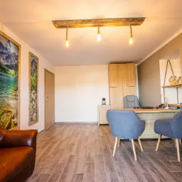 Jasne biuro z drewnianą podłogą, biurkiem z dwoma szarymi krzesłami, brązową skórzaną kanapą i dekoracyjnym oświetleniem sufitowym z żarówkami na sznurkach.