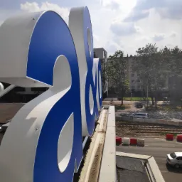 Duże, niebiesko-białe litery na dachu budynku z widokiem na ulicę, tory tramwajowe i budynki mieszkalne w tle.