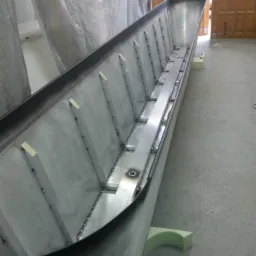 Wnętrze długiego, metalowego koryta z przyspawanymi wzmocnieniami, stojącego na piankowych podporach, widoczne ślady spawania