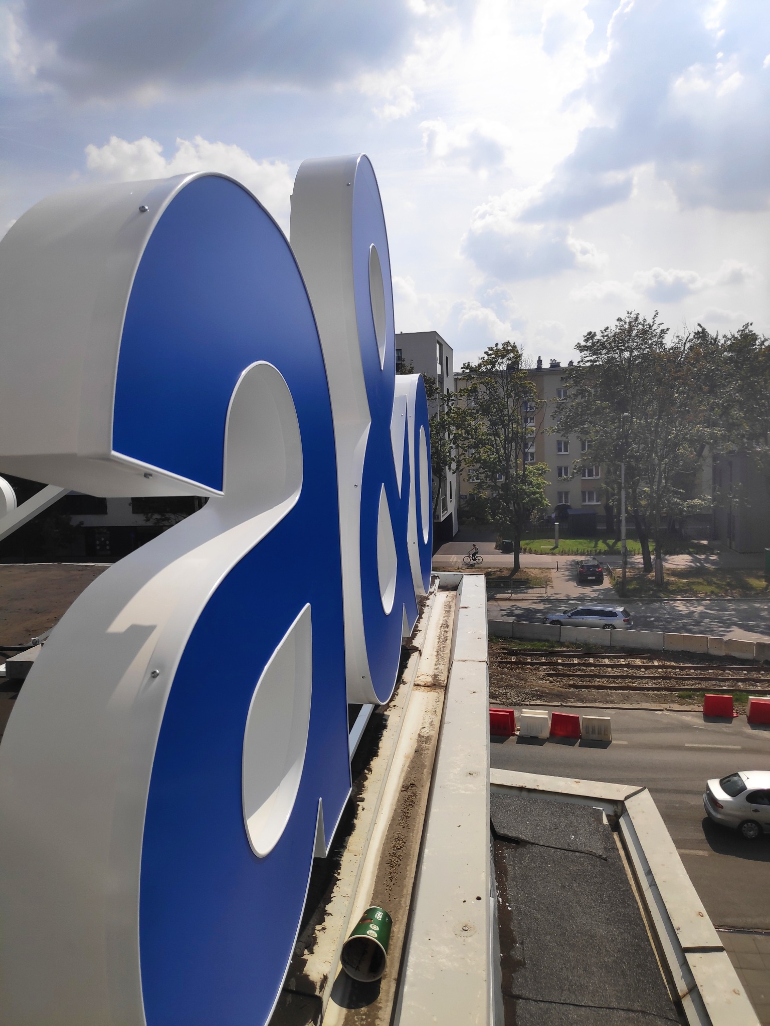 Duże, niebiesko-białe litery na dachu budynku z widokiem na ulicę, tory tramwajowe i budynki mieszkalne w tle.