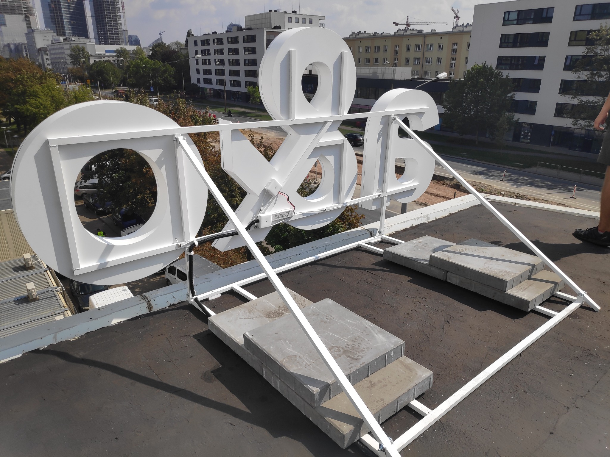Białe, przestrzenne litery i symbole na metalowej konstrukcji, ustawione na dachu budynku z widokiem na panoramę miasta w tle.