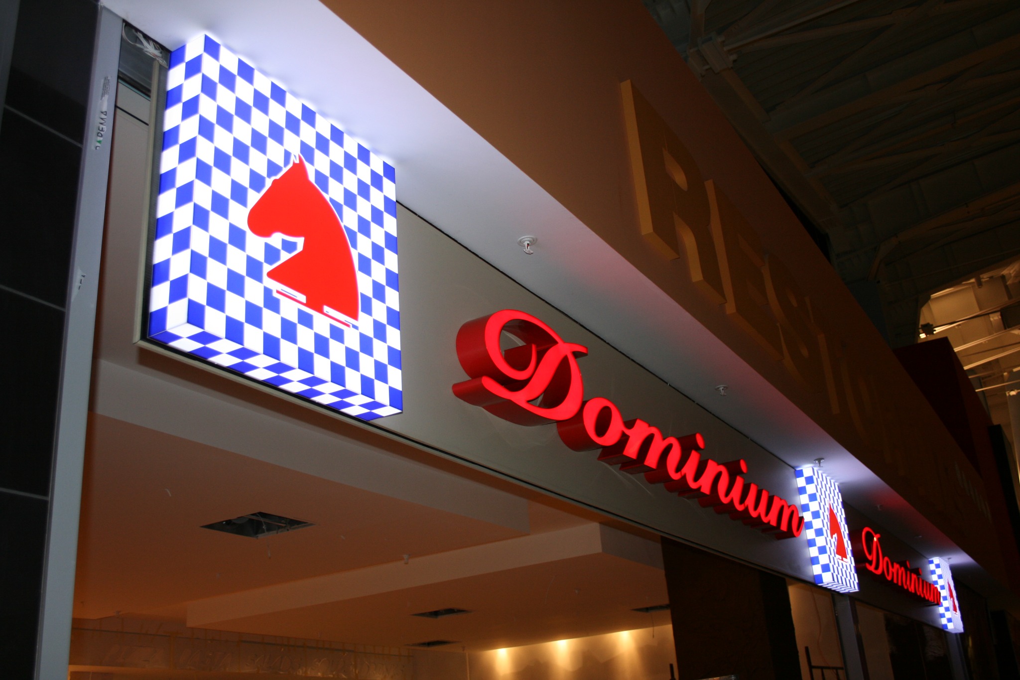 Podświetlany szyld z czerwonym koniem na niebiesko-białej szachownicy i czerwonym napisem 'Dominium' pod sufitem w galerii handlowej.