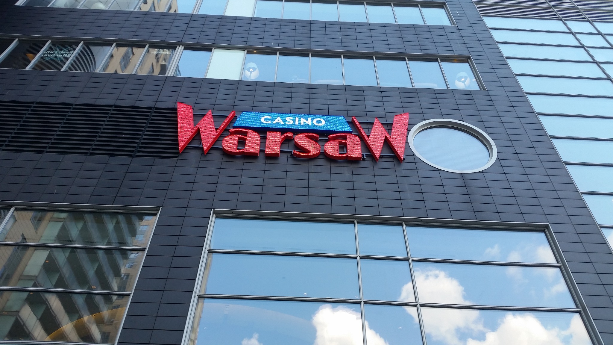 Elewacja budynku z dużym, czerwonym napisem 'Warsaw Casino' podświetlonym na niebiesko, widok z dołu na tle szarej fasady z regularnymi oknami.