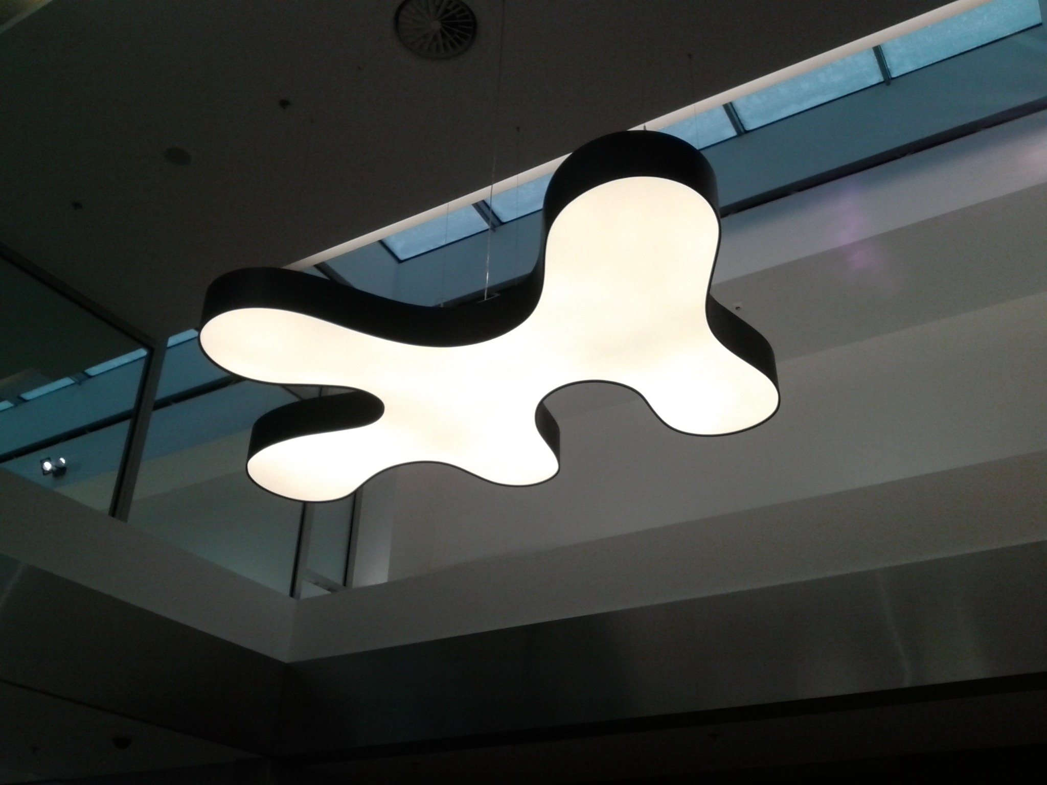 Nowoczesna lampa wisząca o organicznym kształcie, z czarną obudową i białym, rozproszonym światłem, na tle przeszklonego dachu w biurowcu.