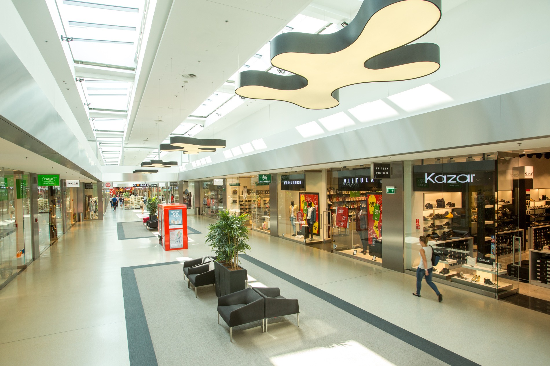 Wnętrze nowoczesnego centrum handlowego z jasną podłogą, przeszklonymi witrynami sklepów i designerskimi lampami sufitowymi w kształcie organicznych form. W tle widoczne świetliki dachowe...