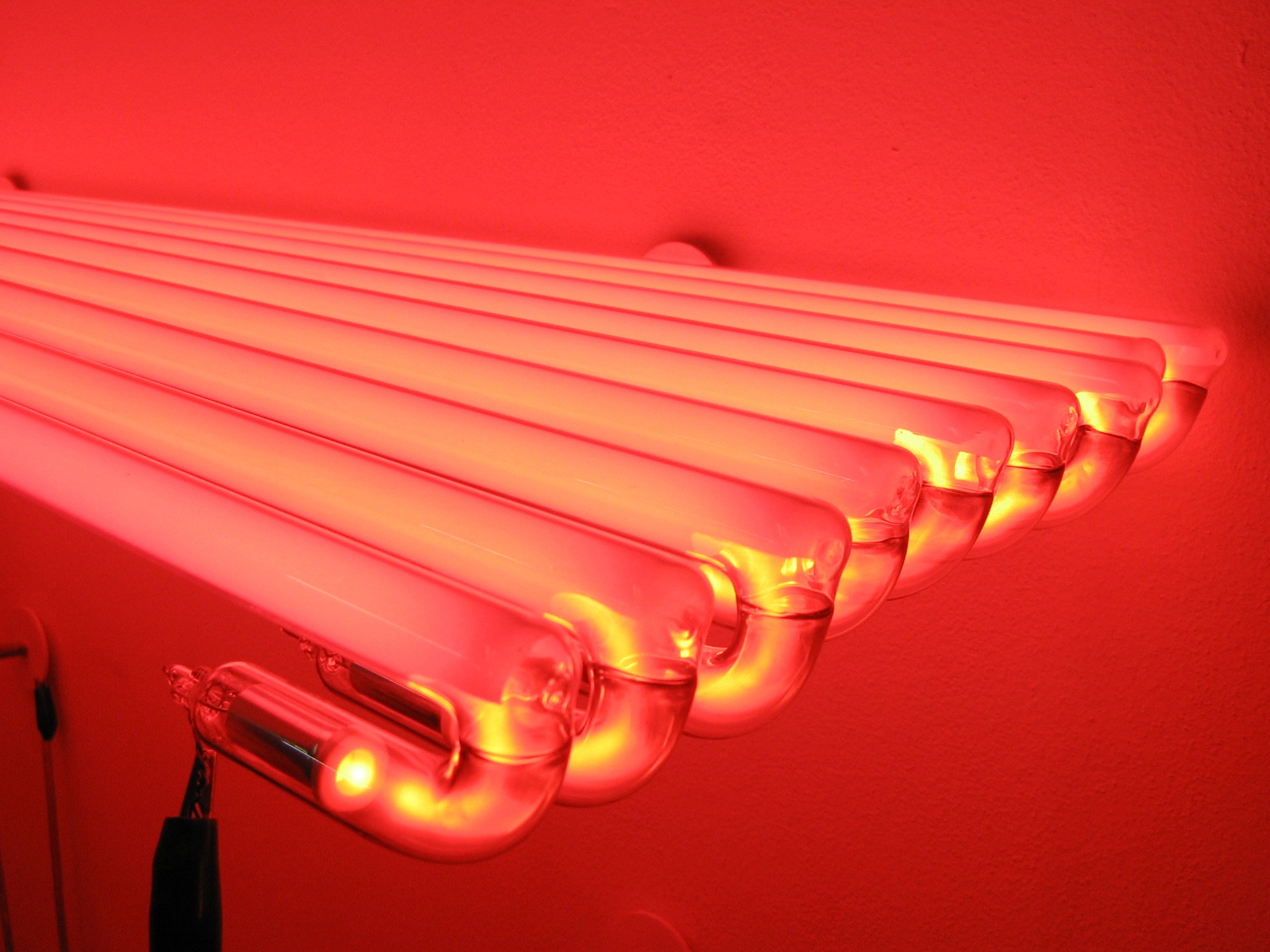 Rząd czerwonych lamp neonowych na ścianie, z widocznymi szklanymi rurami i jasnym światłem. Jedna z lamp ma widoczne przewody.