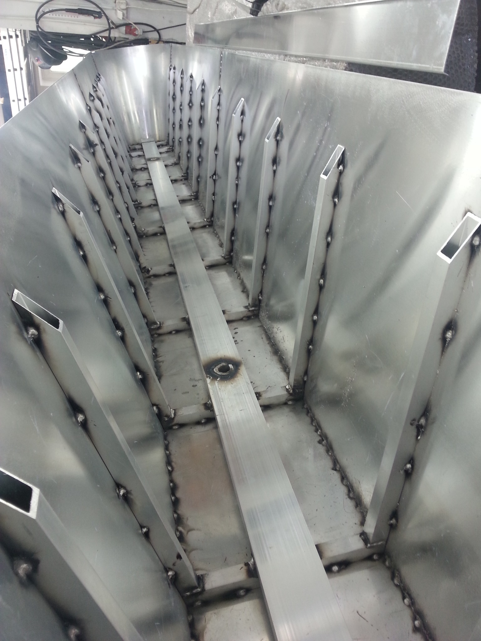 Wnętrze metalowej konstrukcji z licznymi spawami, widoczne metalowe profile o przekroju kwadratowym przymocowane do ścianek, prawdopodobnie etap produkcji.