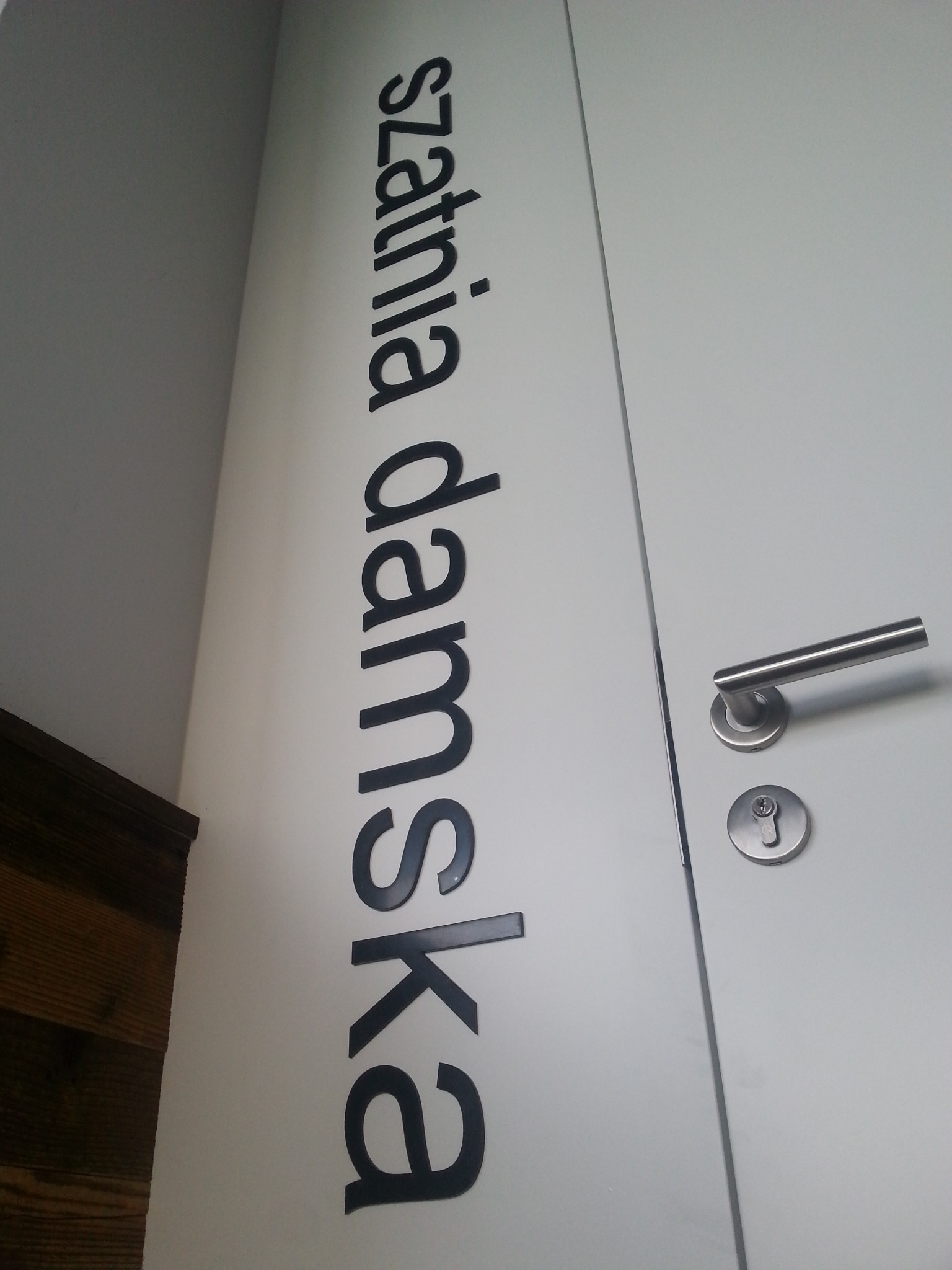 Białe drzwi z metalową klamką i zamkiem, obok pionowy napis 'szatnia damska' wykonany z czarnych liter.