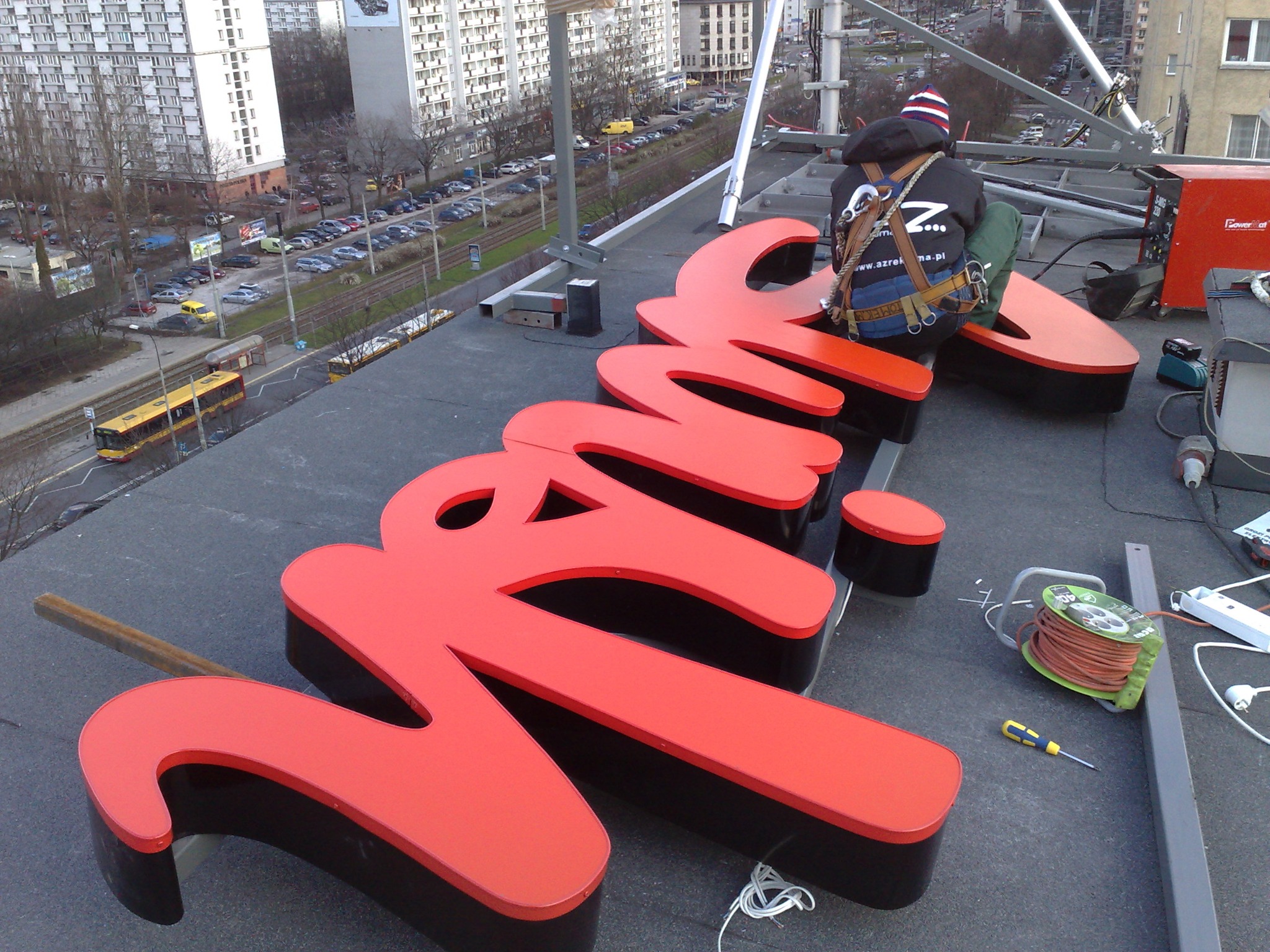 Montaż czerwonego, przestrzennego napisu reklamowego na dachu budynku przez pracownika w uprzęży, z widokiem na panoramę miasta i przejeżdżający tramwaj.