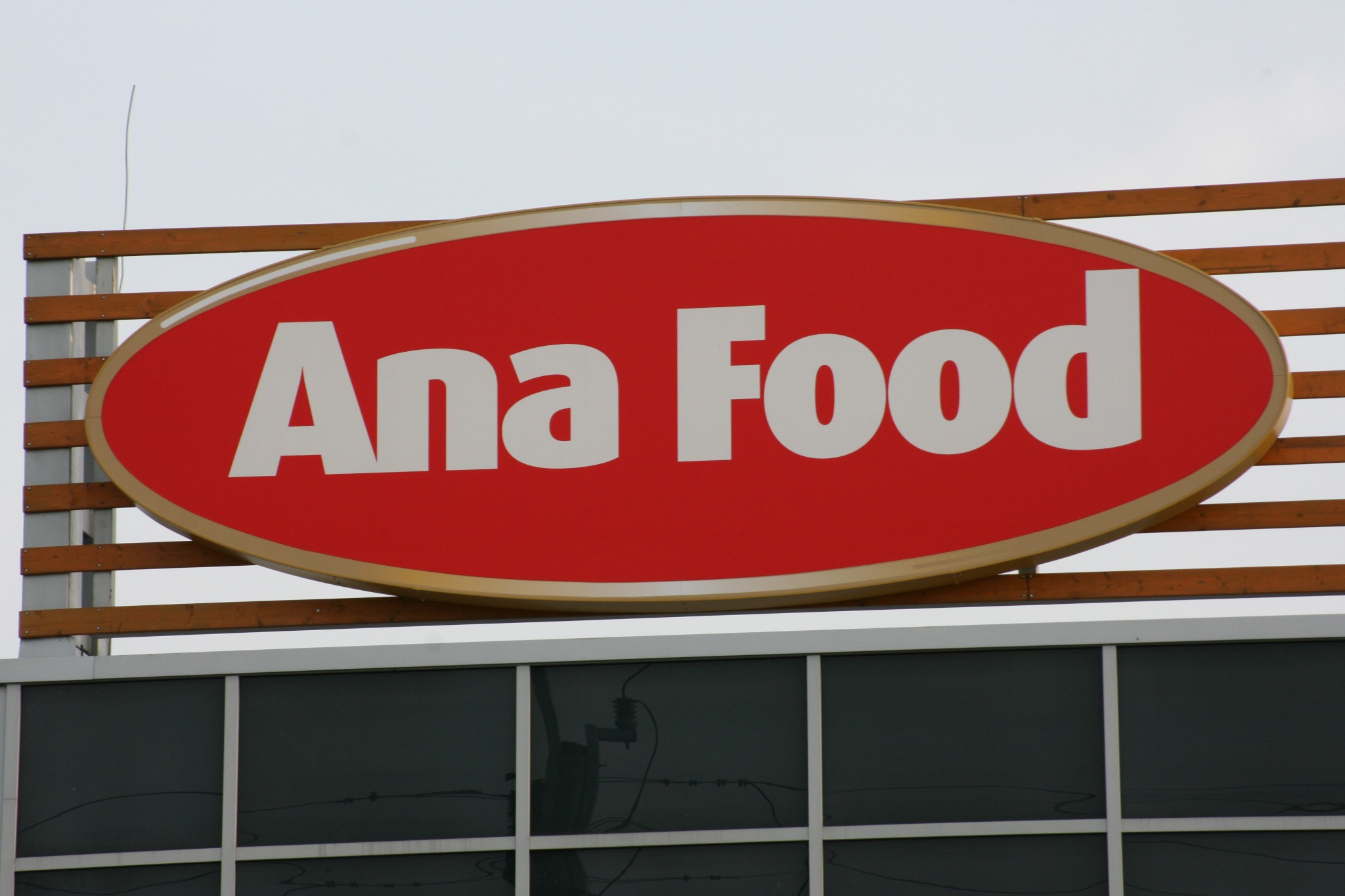 Owalny szyld reklamowy z czerwonym tłem i białym napisem 'Ana Food', umieszczony na drewnianej konstrukcji nad budynkiem z szarymi oknami.