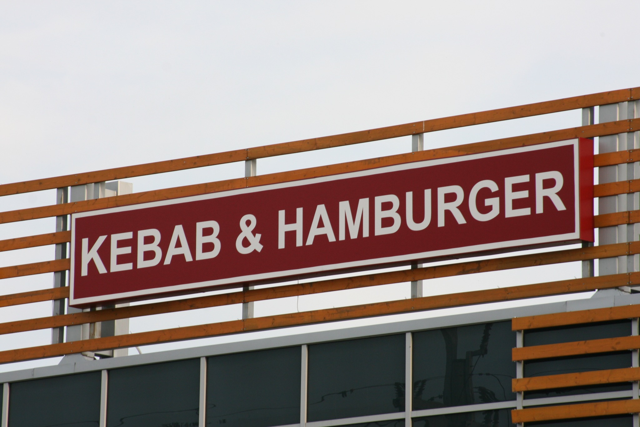 Czerwony szyld z białym napisem 'KEBAB & HAMBURGER' zamontowany na drewnianej konstrukcji nad budynkiem z szarymi oknami.