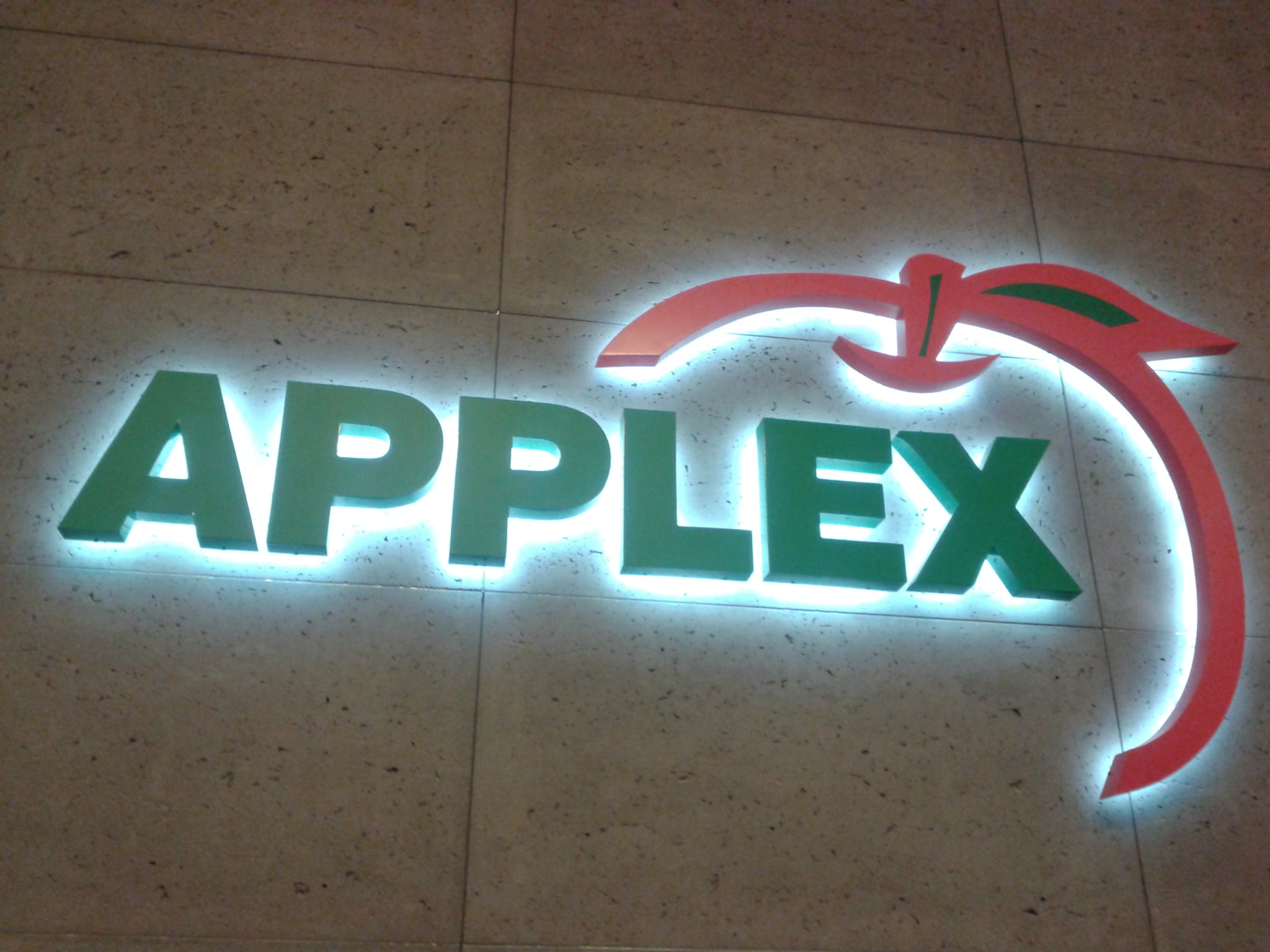 Podświetlane logo firmy APPLex, zielone litery i czerwono-zielony symbol graficzny na tle ściany z jasnych płytek.