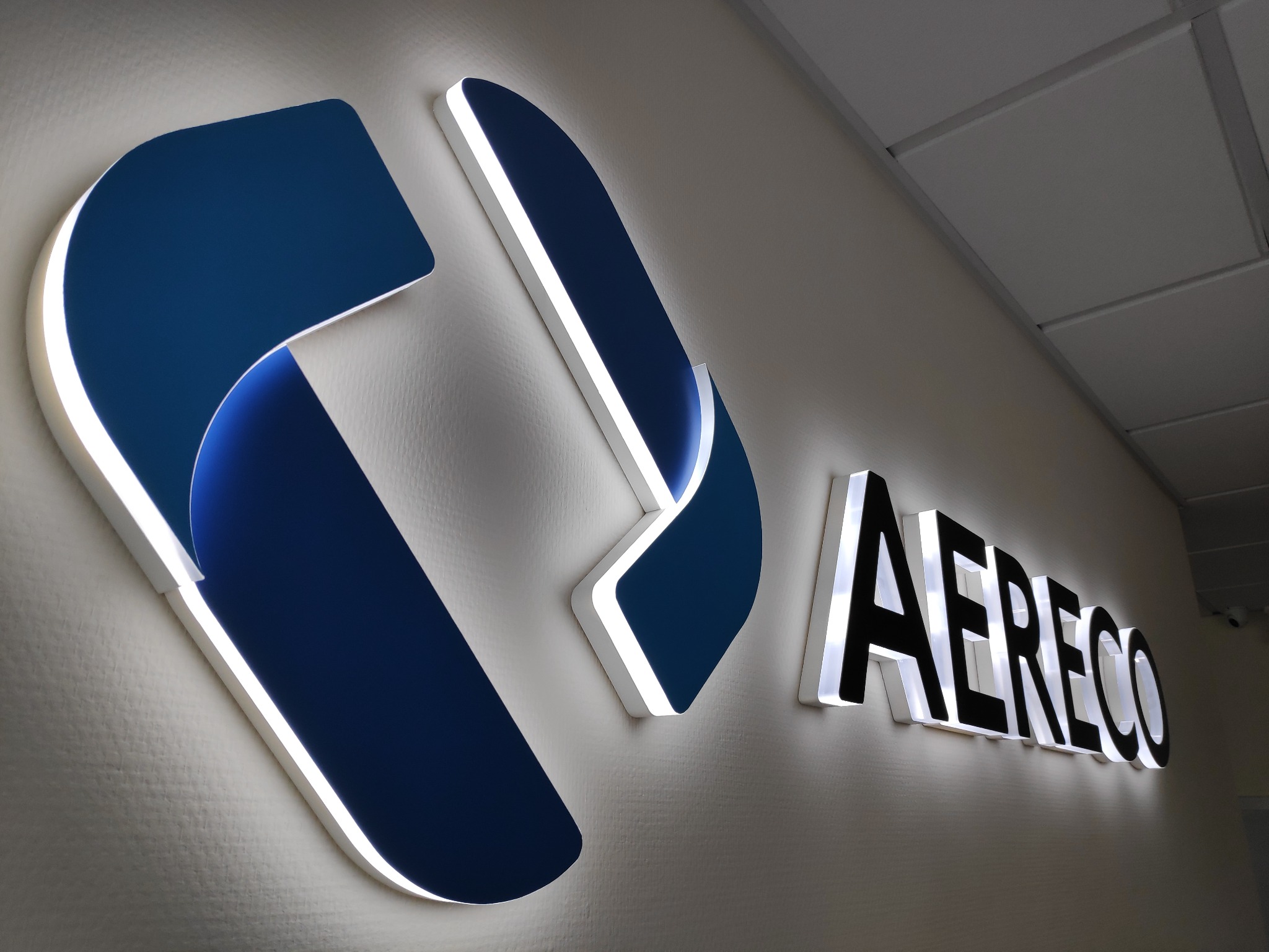 Podświetlane logo firmy AERECO, zamontowane na ścianie w biurze. Logo w kolorze granatowym z białym obramowaniem.