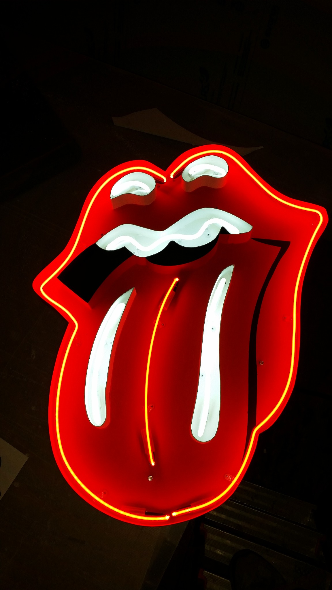 Neonowy znak w kształcie języka i ust, czerwony z żółtą obwódką, oświetlony białymi neonami wewnątrz, na ciemnym tle.