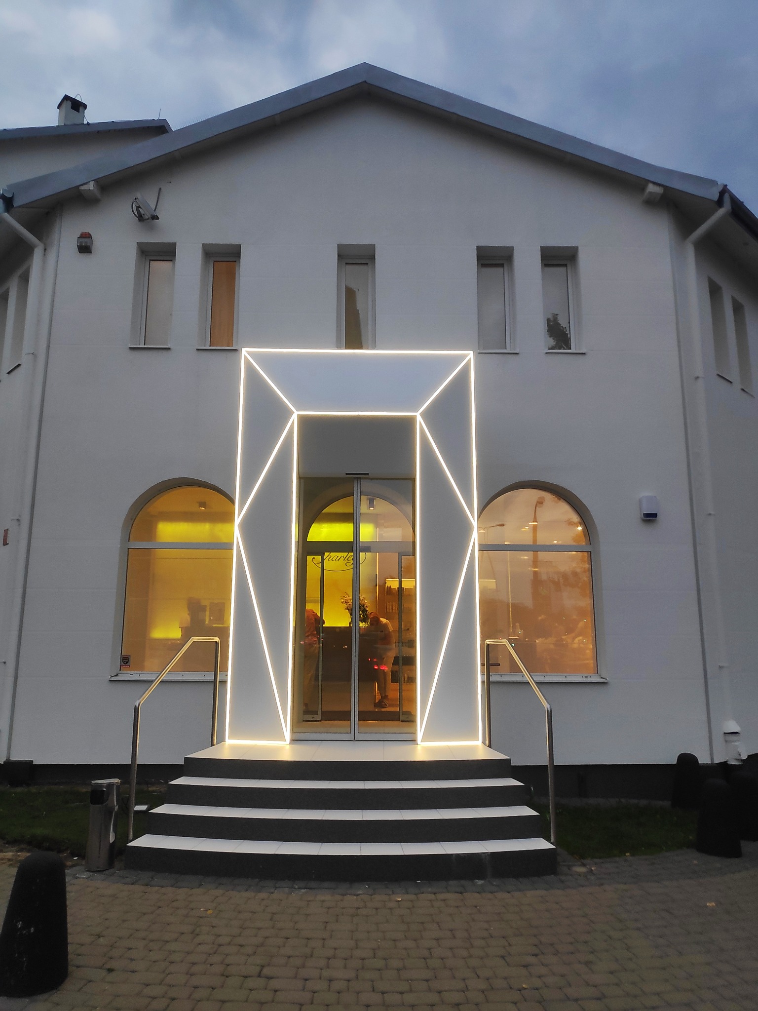Biały budynek z geometryczną, podświetlaną konstrukcją nad wejściem, stalowe balustrady przy schodach, okna łukowe po bokach wejścia.
