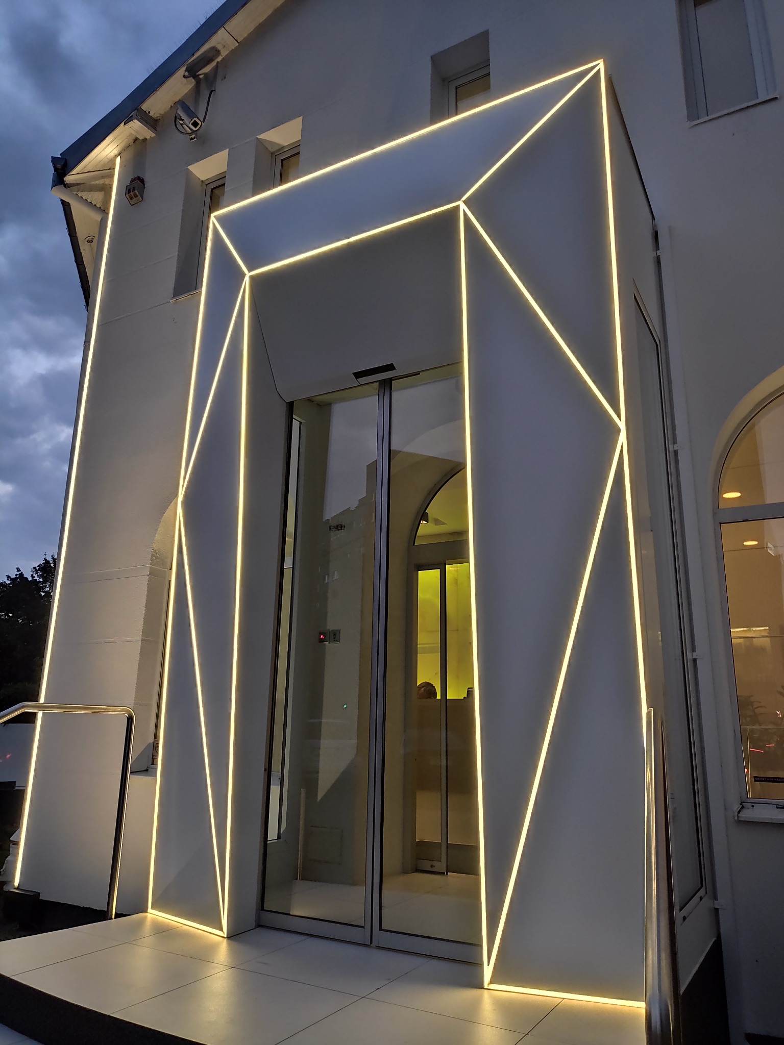 Nowoczesne wejście do budynku z geometrycznym oświetleniem LED w kolorze ciepłej bieli, tworzącym wzór na białej fasadzie wokół szklanych drzwi.