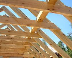 Konstrukcja dachu z jasnego drewna, widoczna od wewnątrz, z fragmentami nieba w prześwitach. Elementy połączone pod kątem, surowe drewno bez wykończenia.