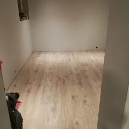 Wykończone wnętrze w Zawadzie z ułożonymi panelami podłogowymi o jasnym, drewnopodobnym wzorze. Ściany przygotowane do dalszych prac, widoczne punkty oświetleniowe w suficie, a w rogu pomieszczenia...