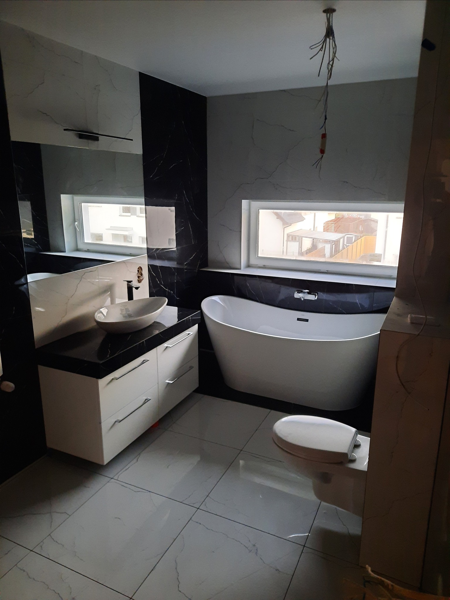 Nowoczesna łazienka w trakcie wykańczania z wolnostojącą wanną, umywalką nablatową na szafce z szufladami, biało-czarne marmurowe płytki na ścianach i niedokończone oświetlenie sufitowe z wiszącymi...