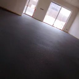 Świeżo wylana, ciemna posadzka w surowym wnętrzu z dwoma oknami balkonowymi, widoczne żółte taśmy zabezpieczające ściany.