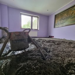 Wylewki Maszynowe z Mixokreta - Pokój z fioletowymi ścianami i oknem, gdzie na podłodze, pokrytej folią, leży gruba warstwa szarego, granulowanego materiału. Na pierwszym planie brudny mieszalnik budowlany.