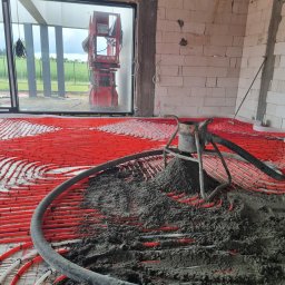 Wylewki Maszynowe z Mixokreta - Układanie jastrychu na czerwonych rurach ogrzewania podłogowego w pomieszczeniu w stanie deweloperskim, z widocznym sprzętem do pompowania i oknem na zewnątrz.