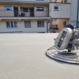 Wylewki Maszynowe z Mixokreta - Zacieraczka mechaniczna marki KREBER na świeżo wylanej, jasnoszarej posadzce o drobnoziarnistej strukturze, z blokiem mieszkalnym i balkonami w tle, w słoneczny dzień.