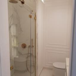 Fhu Jaroszewicz - Minimalistyczna łazienka w bieli z akcentami złota: kabina prysznicowa ze szklanymi drzwiami, marmurowe płytki, armatura w kolorze złota, podwieszana toaleta i oświetlenie LED.