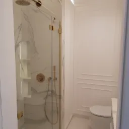 Minimalistyczna łazienka w bieli z akcentami złota: kabina prysznicowa ze szklanymi drzwiami, marmurowe płytki, armatura w kolorze złota, podwieszana toaleta i oświetlenie LED.