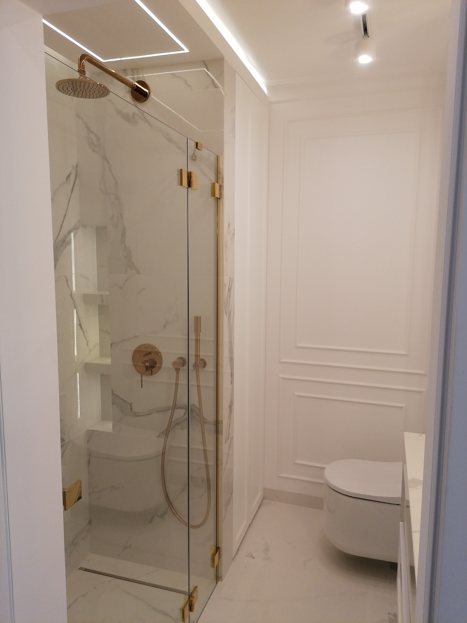 Minimalistyczna łazienka w bieli z akcentami złota: kabina prysznicowa ze szklanymi drzwiami, marmurowe płytki, armatura w kolorze złota, podwieszana toaleta i oświetlenie LED.