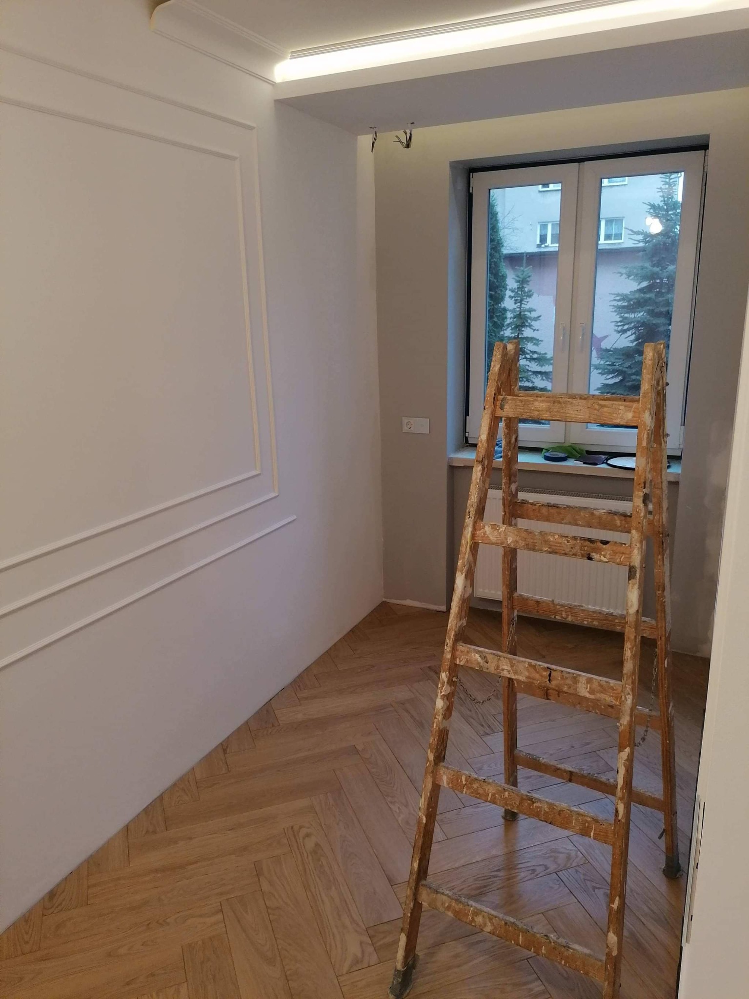 Świeżo pomalowane wnętrze z sztukaterią na ścianie, drewnianą podłogą w jodełkę i starą, zabrudzoną farbą drabiną opartą o ścianę przy oknie.