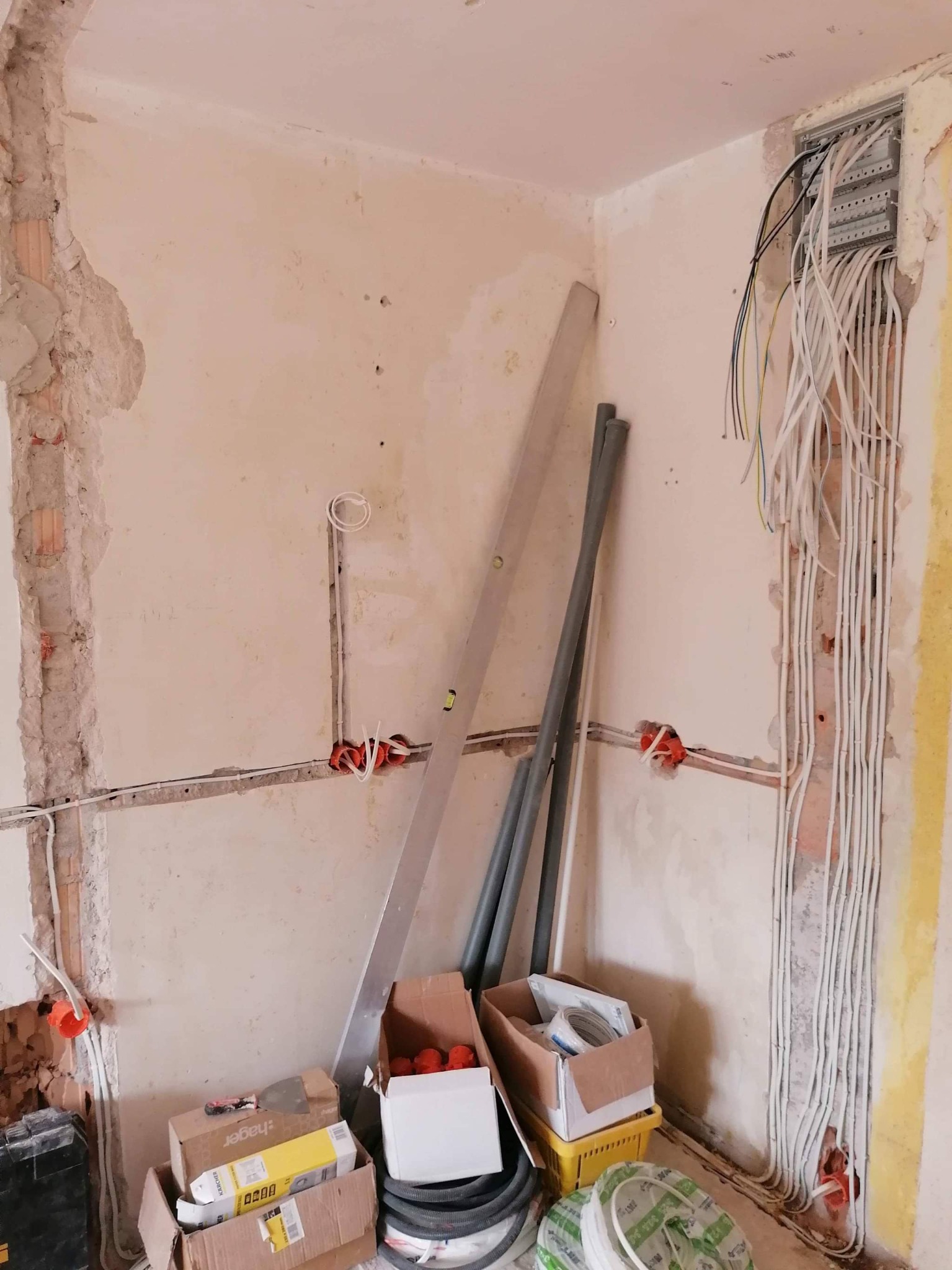 Ściana w trakcie prac elektrycznych: widoczne kable w bruzdach, puszki instalacyjne, skrzynka rozdzielcza z licznymi przewodami, poziomica oparta o ścianę oraz kartony z akcesoriami elektrycznymi.