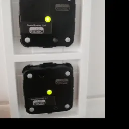 Dwa czarne, kwadratowe czujniki alarmowe z żółtymi diodami LED, zamontowane w białej ramce na ścianie.