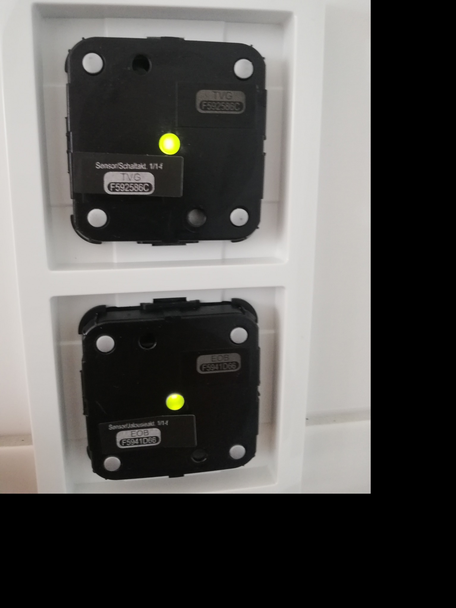 Dwa czarne, kwadratowe czujniki alarmowe z żółtymi diodami LED, zamontowane w białej ramce na ścianie.